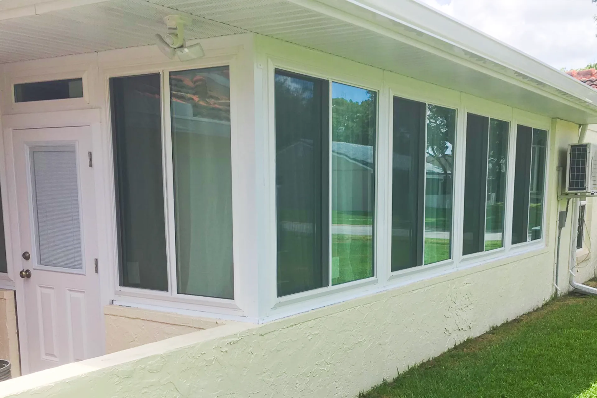 A white-framed sunroom running along the side of a home, with a long run of windows, an entry door and a wall-mounted air handler